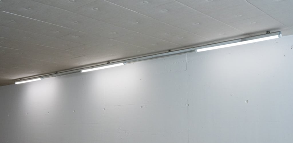 Das Bild zeigt eine helle Wand- und Deckeninstallation mit linear angeordneten TRIVALITE Perseus Leuchten. Die Beleuchtung sorgt für gleichmässiges, blendfreies Licht und eignet sich ideal für Durchgänge, Zufahrten oder Tiefgaragen. TRIVALITE Perseus Wandleuchte im Tunnelbereich
