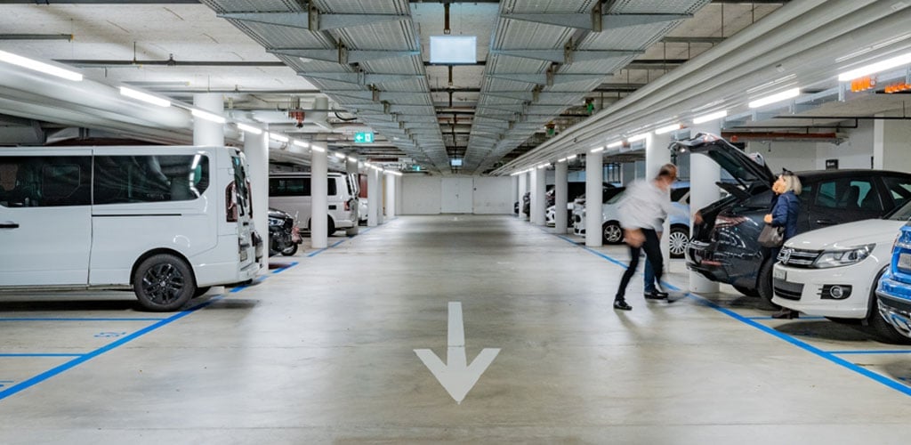 Das Bild zeigt ein modernes Parkhaus mit klar strukturierter Fahrgasse und Fahrzeugen beidseitig. Die Decke ist mit dem TRIVALITE Perseus Schienensystem ausgestattet, das für eine gleichmässige, energieeffiziente Ausleuchtung sorgt. Eine grosse Pfeilmarkierung weist die Fahrtrichtung. Parkhaus mit TRIVALITE Perseus Lichtsystem