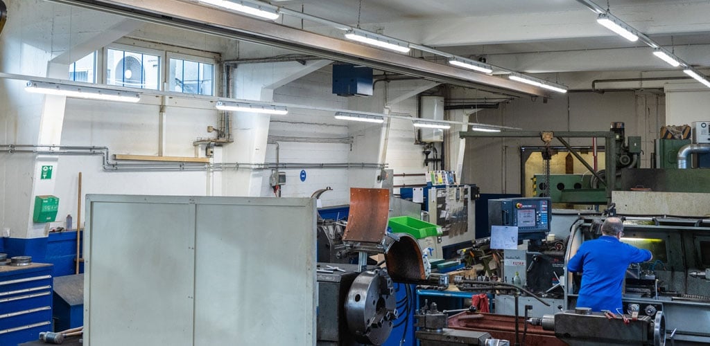 Das Bild zeigt eine industrielle Werkstatt mit Maschinen und Werkzeugen. An der Decke sind mehrere TRIVALITE Perseus Leuchten installiert, die eine helle, gleichmässige Ausleuchtung des Arbeitsbereichs gewährleisten – ideal für präzises Arbeiten und energieeffiziente Beleuchtung in Produktionsumgebungen. Werkstatt mit TRIVALITE Perseus Beleuchtungssystem