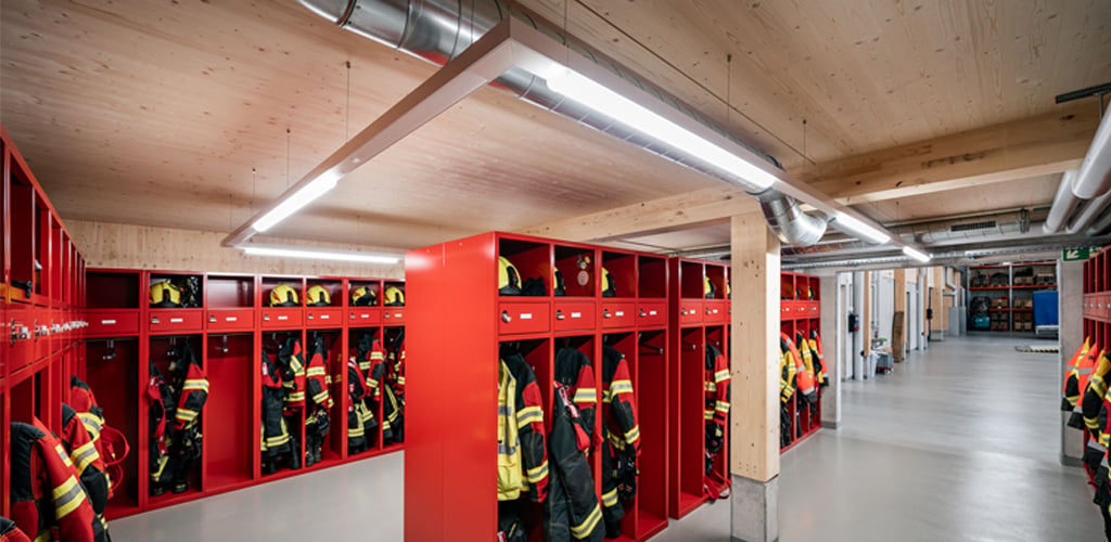 Das Bild zeigt den Umkleidebereich einer Feuerwehr mit roten Spinden und Einsatzkleidung. Über den Reihen sind TRIVALITE Perseus Leuchten installiert, die für eine gleichmässige, helle Beleuchtung sorgen. Die Installation fügt sich harmonisch in die Holzdecke und das industrielle Design ein. Feuerwehrgarderobe mit TRIVALITE Perseus Beleuchtung
