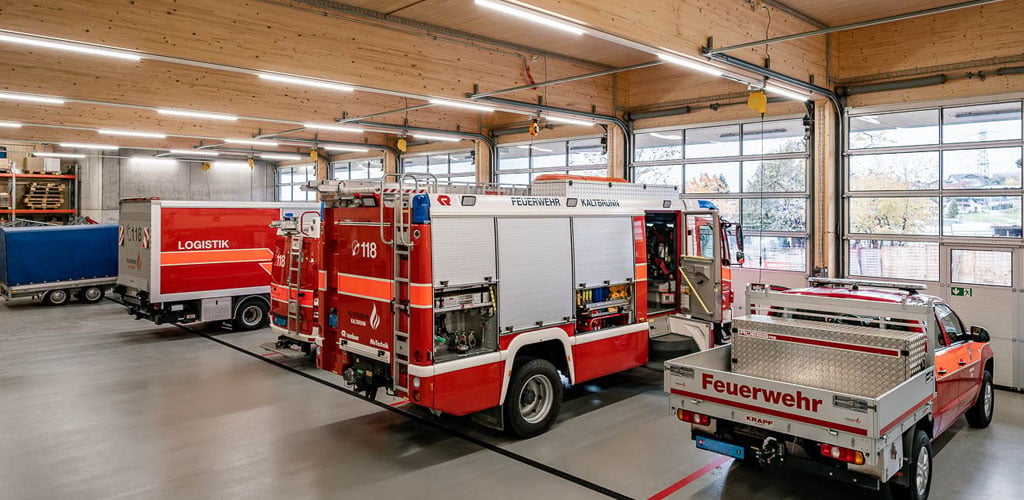 Das Bild zeigt eine moderne Fahrzeughalle der Feuerwehr mit mehreren Einsatzfahrzeugen. Die Decke aus Holz ist mit dem TRIVALITE Perseus Schienensystem ausgestattet, das für eine helle, gleichmässige und energieeffiziente Ausleuchtung der gesamten Halle sorgt. Feuerwehrdepot mit TRIVALITE Perseus Beleuchtungssystem