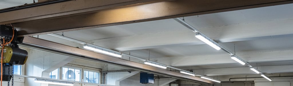 In einer hohen Industriehalle hängen mehrere lineare LED-Leuchten an Stahlträgern und sorgen für eine helle, gleichmässige Grundbeleuchtung. Industriehalle mit mehreren LED-Leuchten an der Decke.