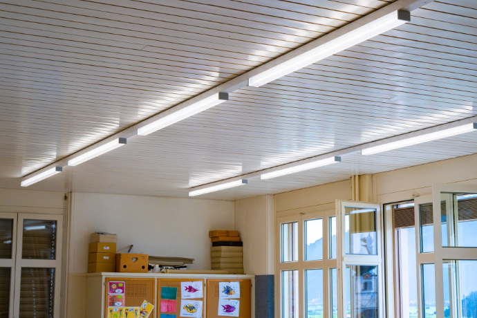 Zwei längliche LED-Leuchten sind an einer hellen, lamellenförmigen Metalldecke montiert und sorgen für eine gleichmässige, moderne Ausleuchtung. Deckenansicht mit zwei linearen LED-Leuchten auf einer lamellenartigen Metallverkleidung.