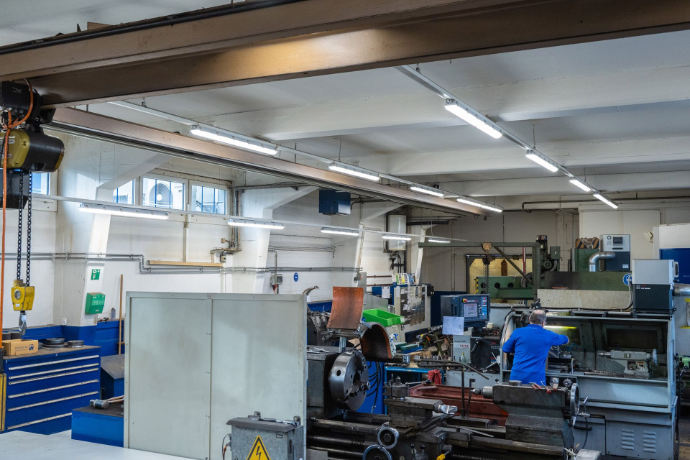 In einer hohen Industriehalle hängen mehrere lineare LED-Leuchten an Stahlträgern und sorgen für eine helle, gleichmässige Grundbeleuchtung. Industriehalle mit mehreren LED-Leuchten an der Decke.
